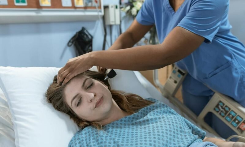 Crop African American female doctor with professional equipment doing examination of ear of woman lying on bed in hospital ward